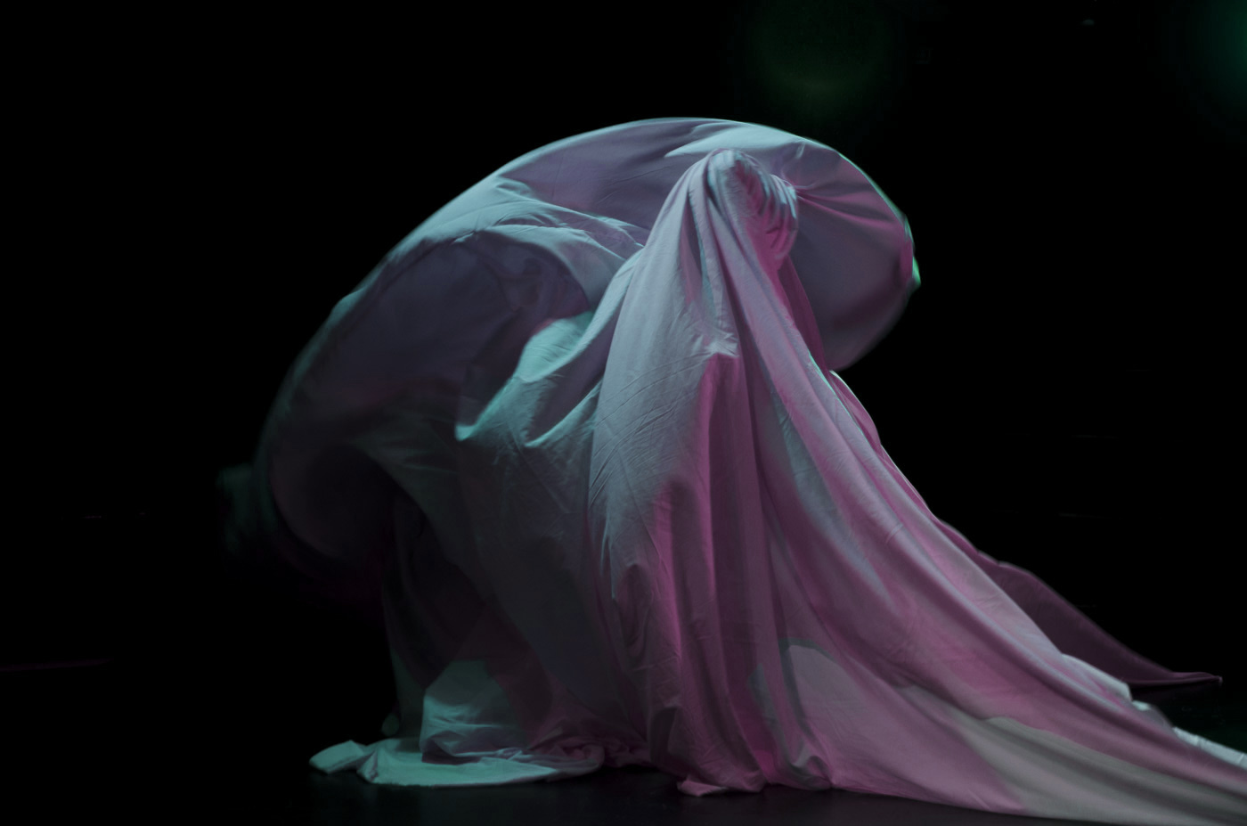 A bluish-green and pink lit fabric that appears to be captured in the middle of some sort of movement. There is a sense of spiraling and twisting of the fabric.