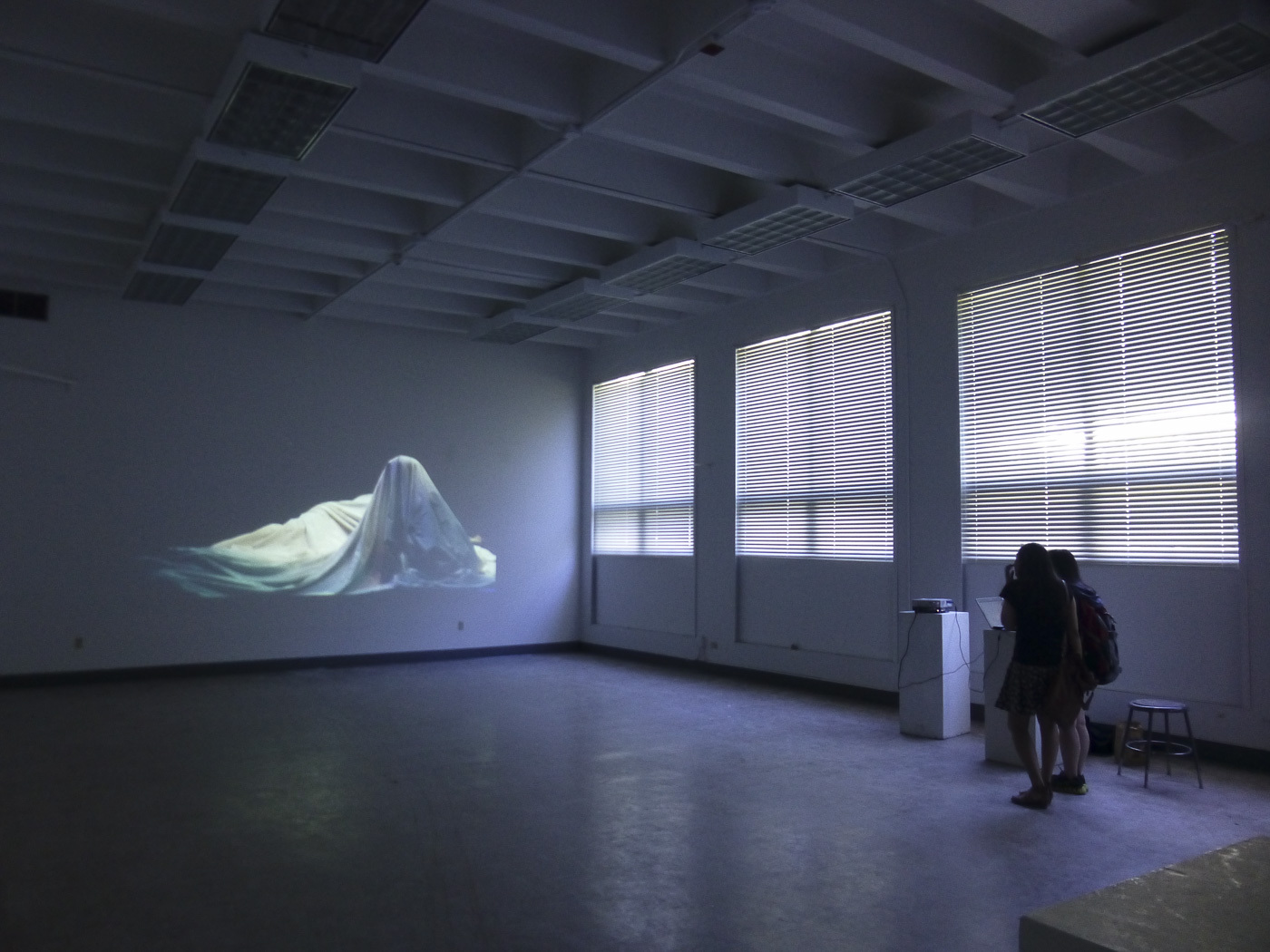 two people stand at one end of a large empty room while a video projection of a figure underneath a large sheet of fabric is displayed on the opposite wall