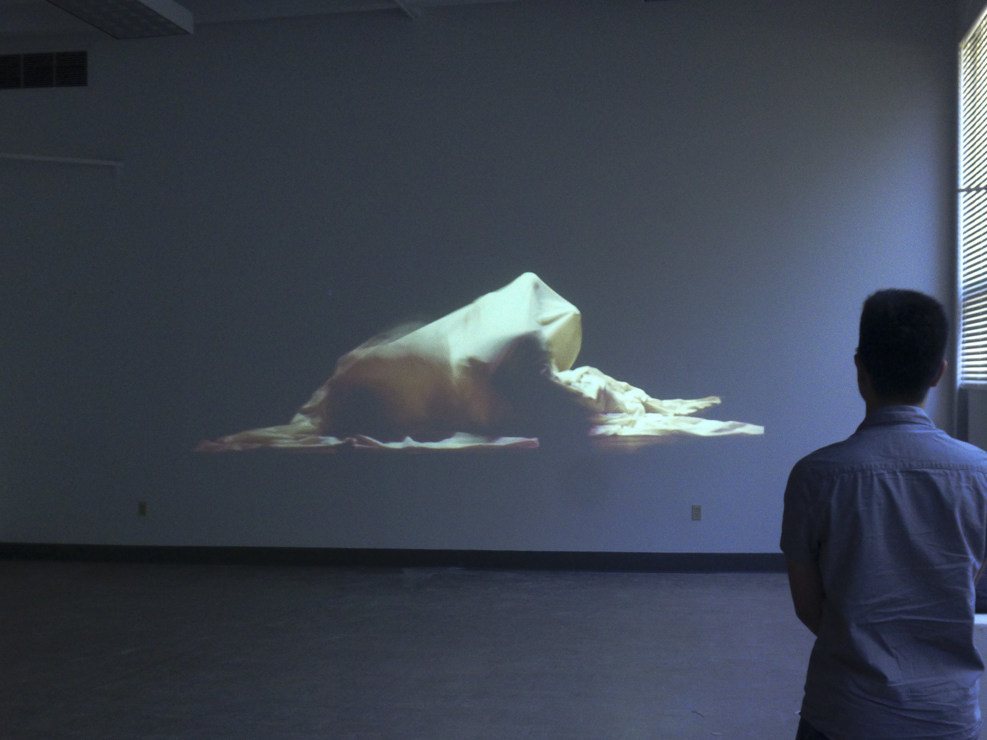 one person stands watching a video projection of an abstract fabric shape displayed on the wall of a dark empty room