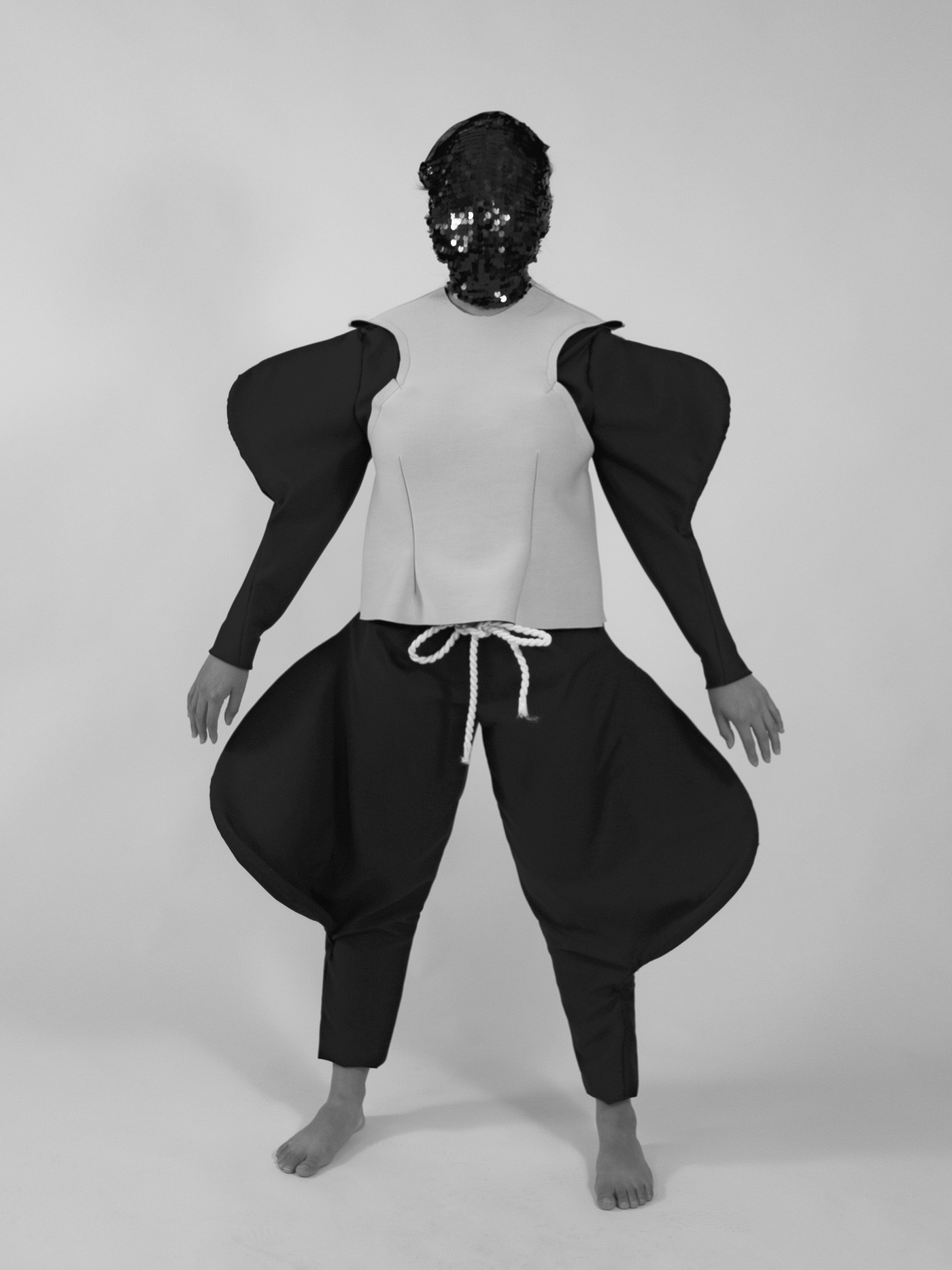 A figure wearing a sequin-covered mask wears a bodysuit with bulging shapes at the thigh and bicep area. A white rope ties the pants together.