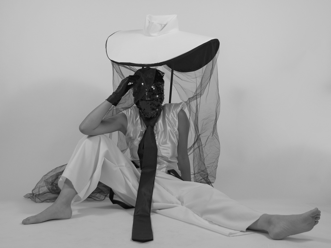 A black and white image of a figure seated on the ground while wearing a sequin-covered mask. They also wear a long black tie, black gloves, white pants, and a hat which has a black mesh-like fabric falling from the brim.