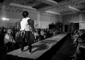 A black and white image of a figure at the end of a fashion runway with an audience of people on either side of it. The model's face is obscured by a sequin covered mask and a wears a black and white outfit that exaggerates the bicep and thigh muscle areas of the model.
