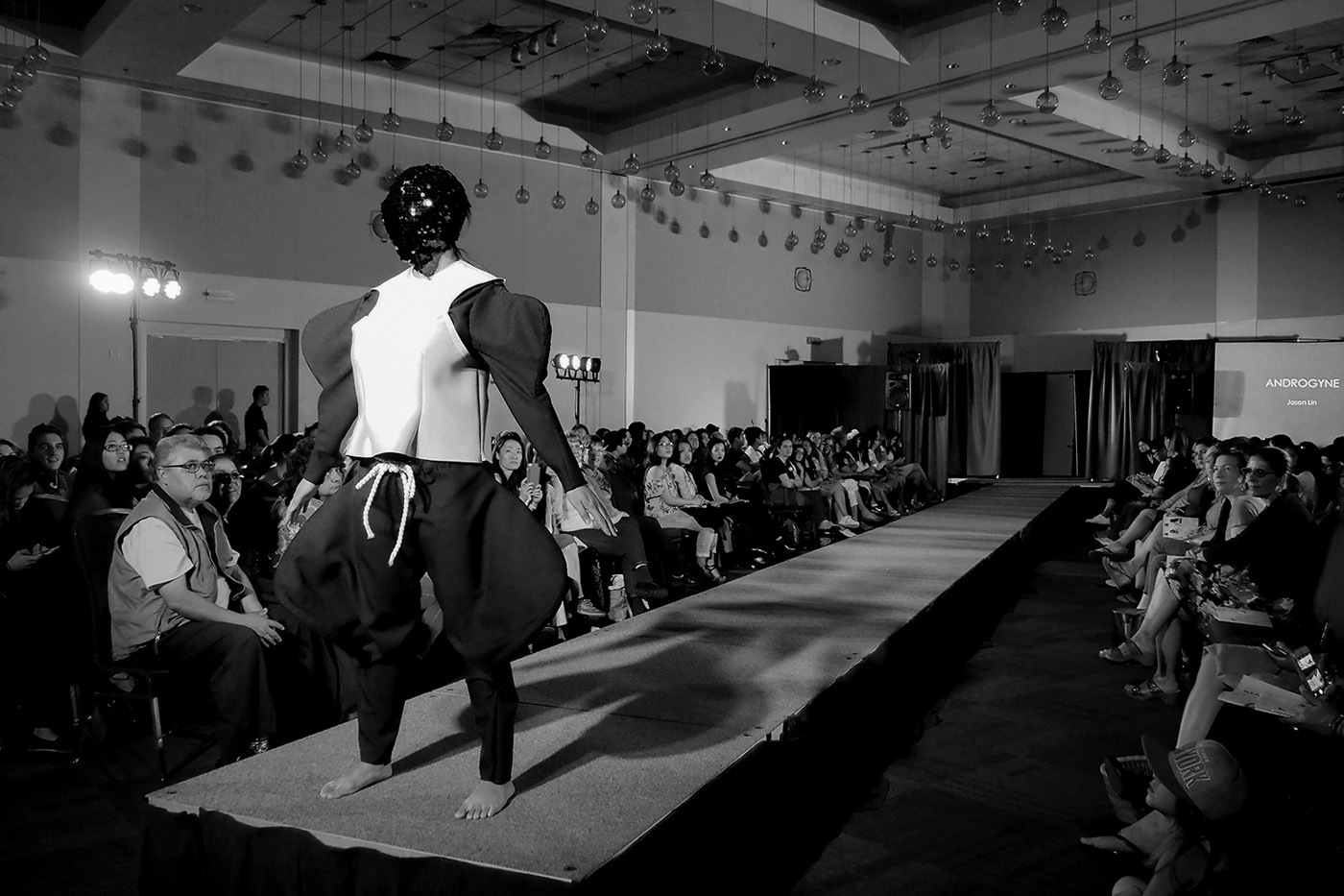 A black and white image of a figure at the end of a fashion runway with an audience of people on either side of it. The model's face is obscured by a sequin covered mask and a wears a black and white outfit that exaggerates the bicep and thigh muscle areas of the model.