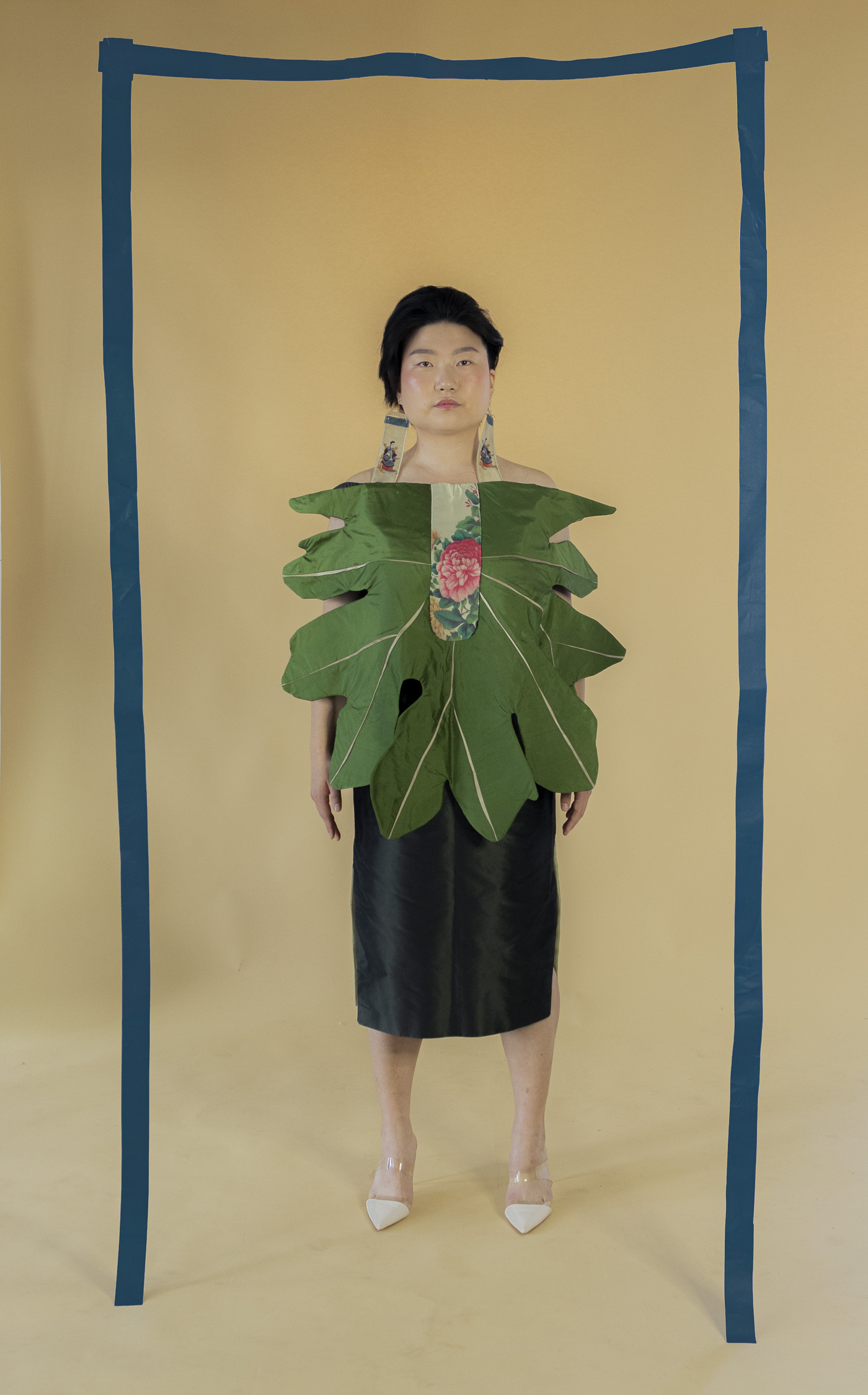 Dahn Bi stands wearing a leaf inspired green dress. They are framed with hanging blue tape