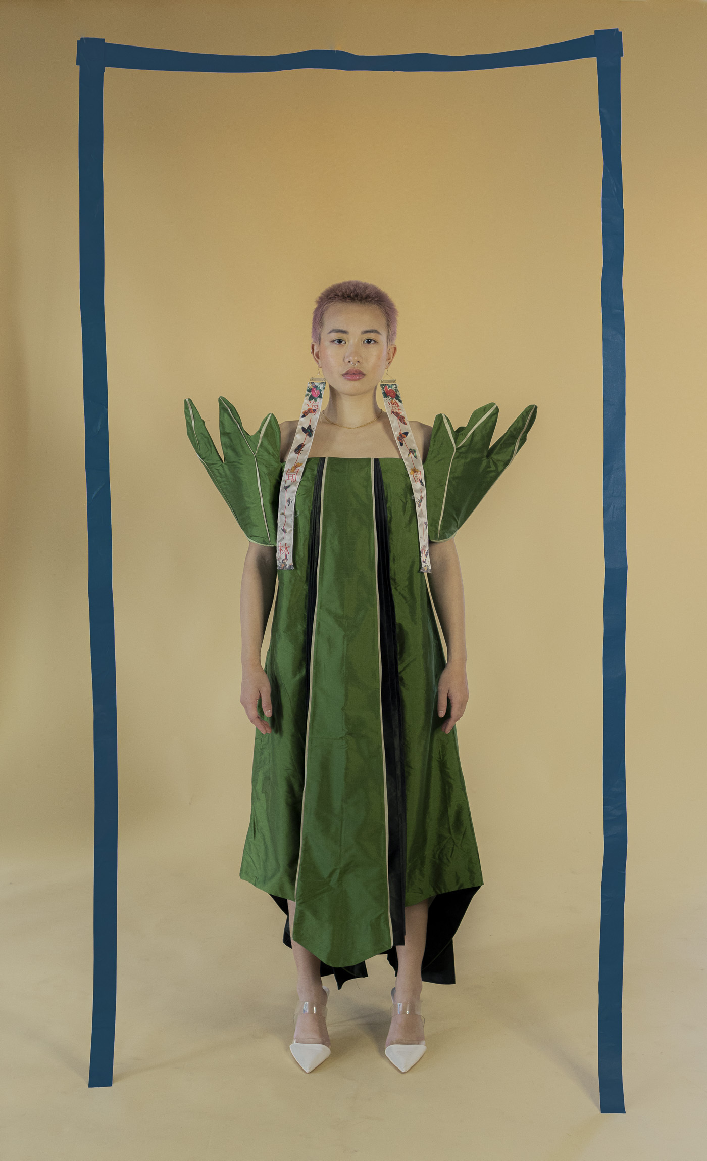 Lucky stands wearing a leaf inspired green and white dress. They are framed with hanging blue tape