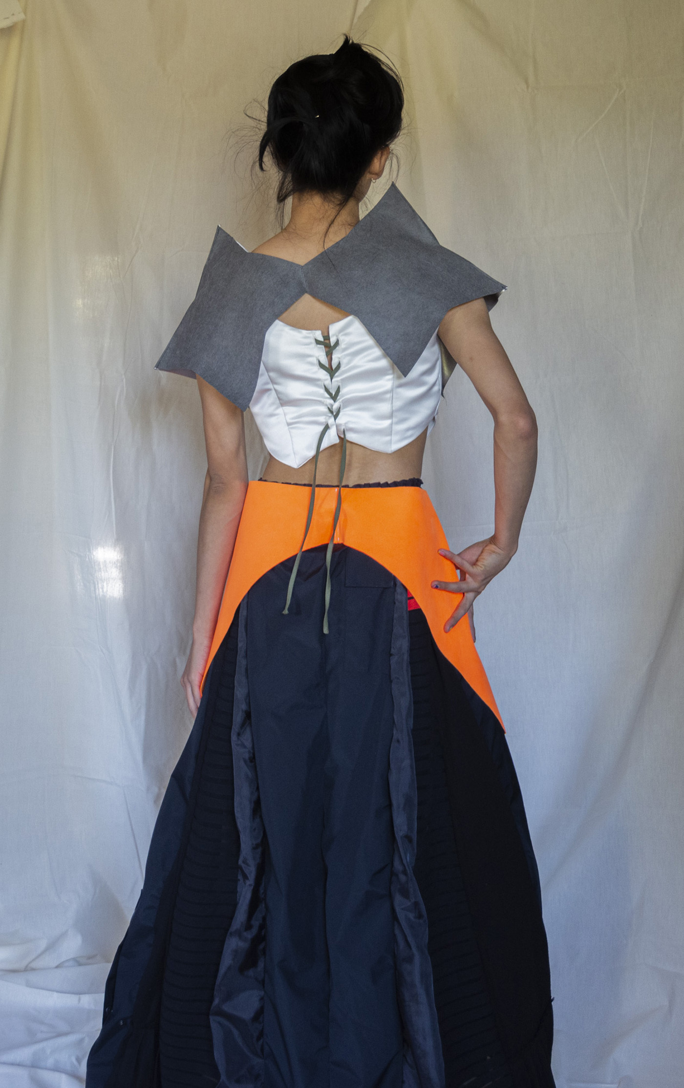 backside view of the Project Upcycle garment. Two four-pointed star shaped shoulder pieces stand over a white shiny corset piece that is tied at the back with green fabric. The dress is black with a bright neon orange geometric shape on top.