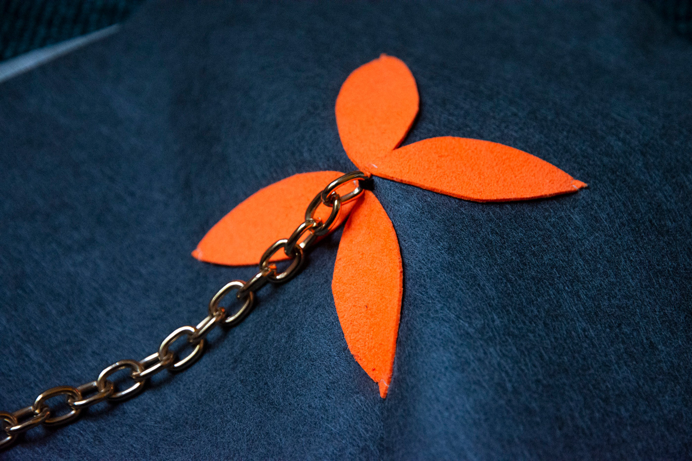A flower-shaped neon orange motif is sewn on top of a matted grey fabric. From the center of the motif, a long gold chain is attached.