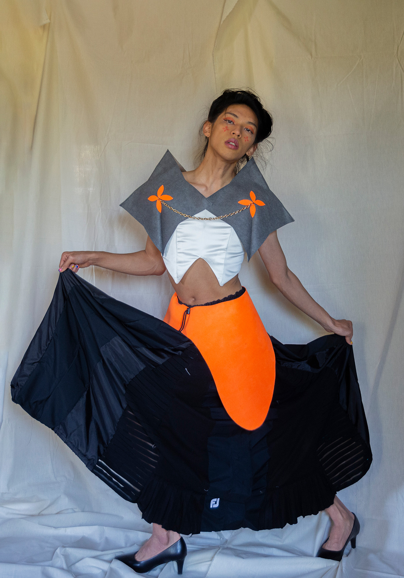 A model poses with a dress. They are holding up two sides of the dress revealing heels. A bright orange piece sits on top of the black dress.