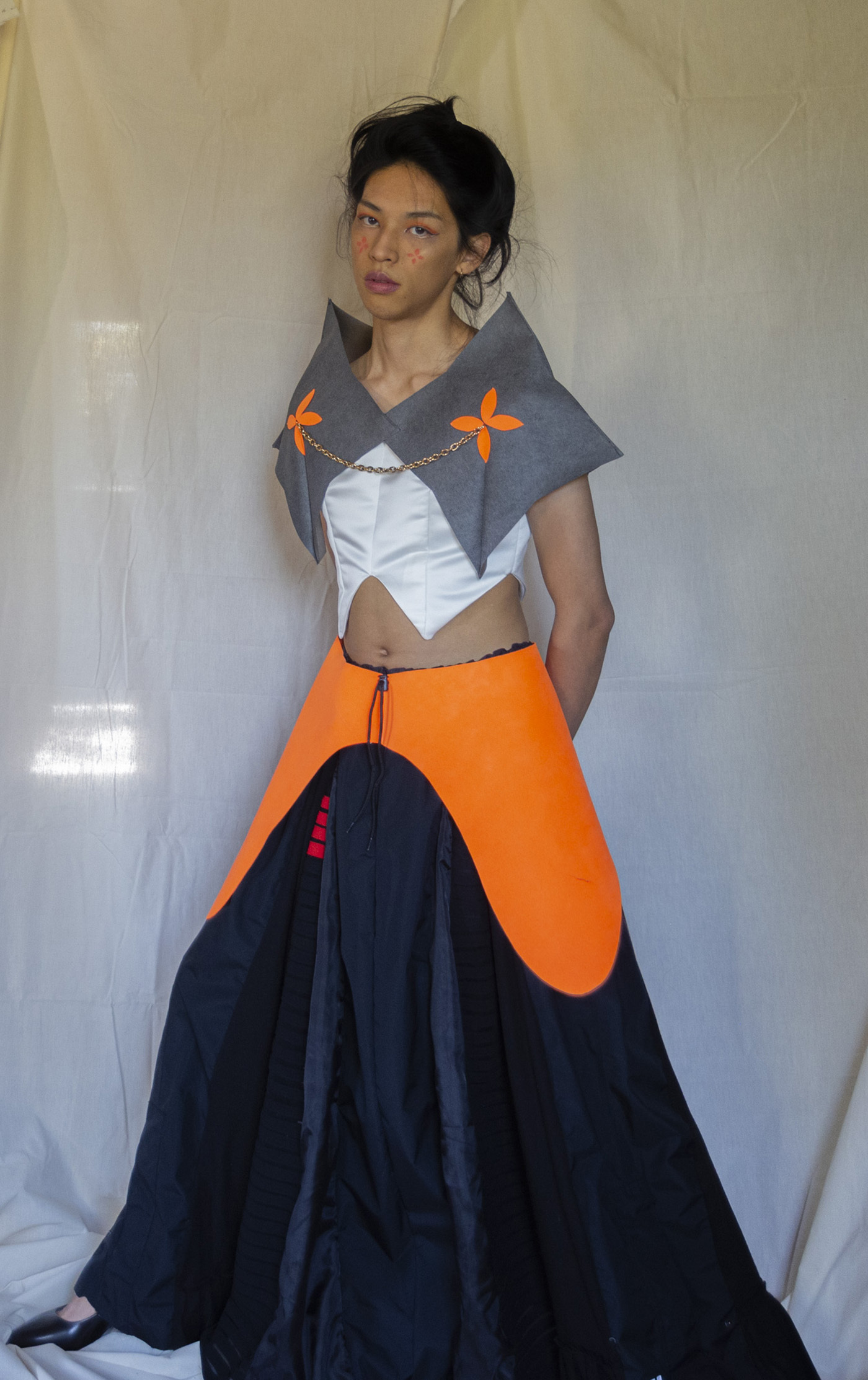 Front view of the Project Upcycle garment. There are two large four-pointed star-shaped shoulder pieces with four-pointed petal-like shapes in the middle of them. There is a bright neon orange geometric bottom piece on top of a long flowing black dress.