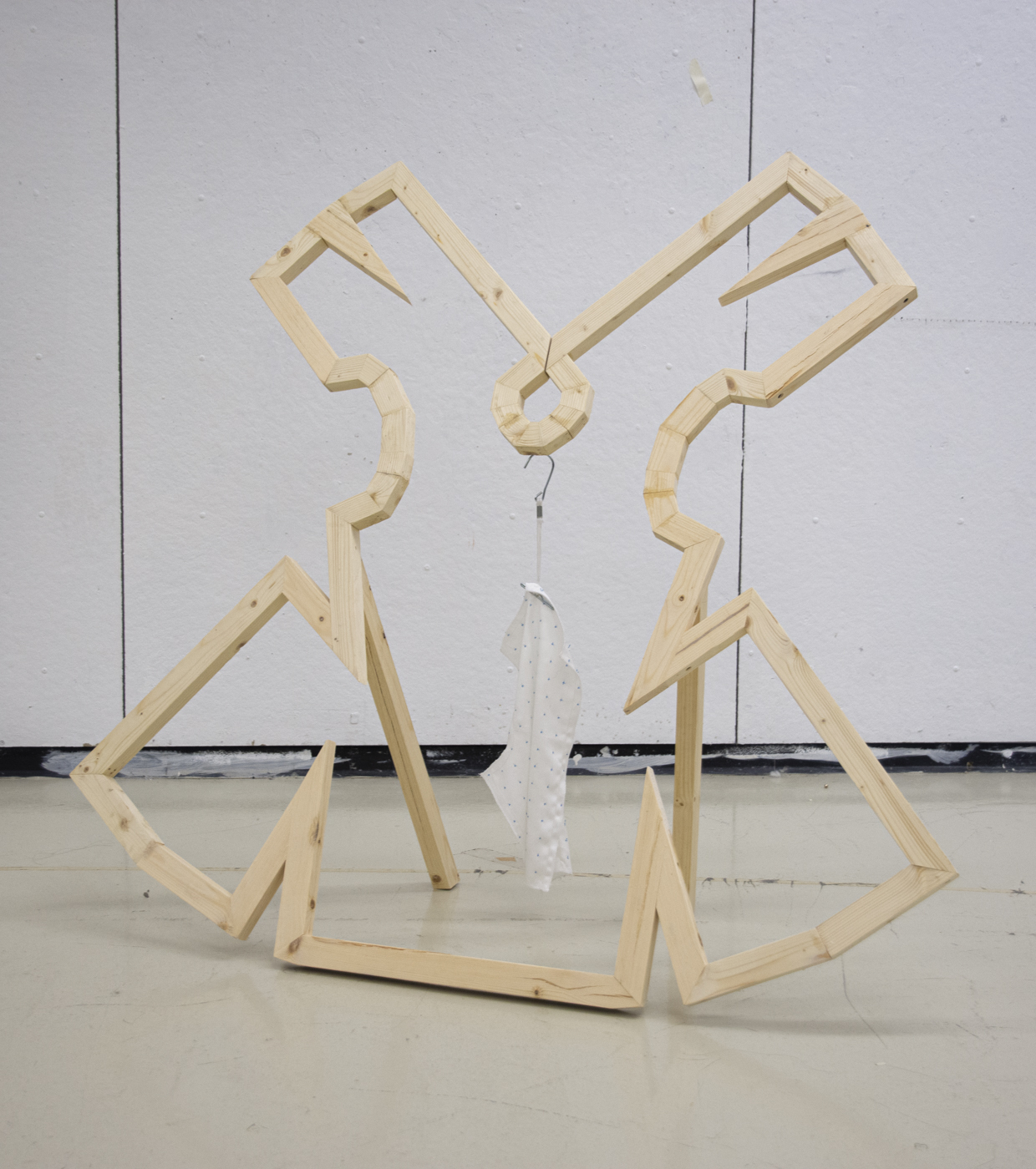 a wooden frame in an abstract shape leans back on two wooden legs. From the center of the shape, a pattern hook hangs. A white fabric hangs from the patternhook