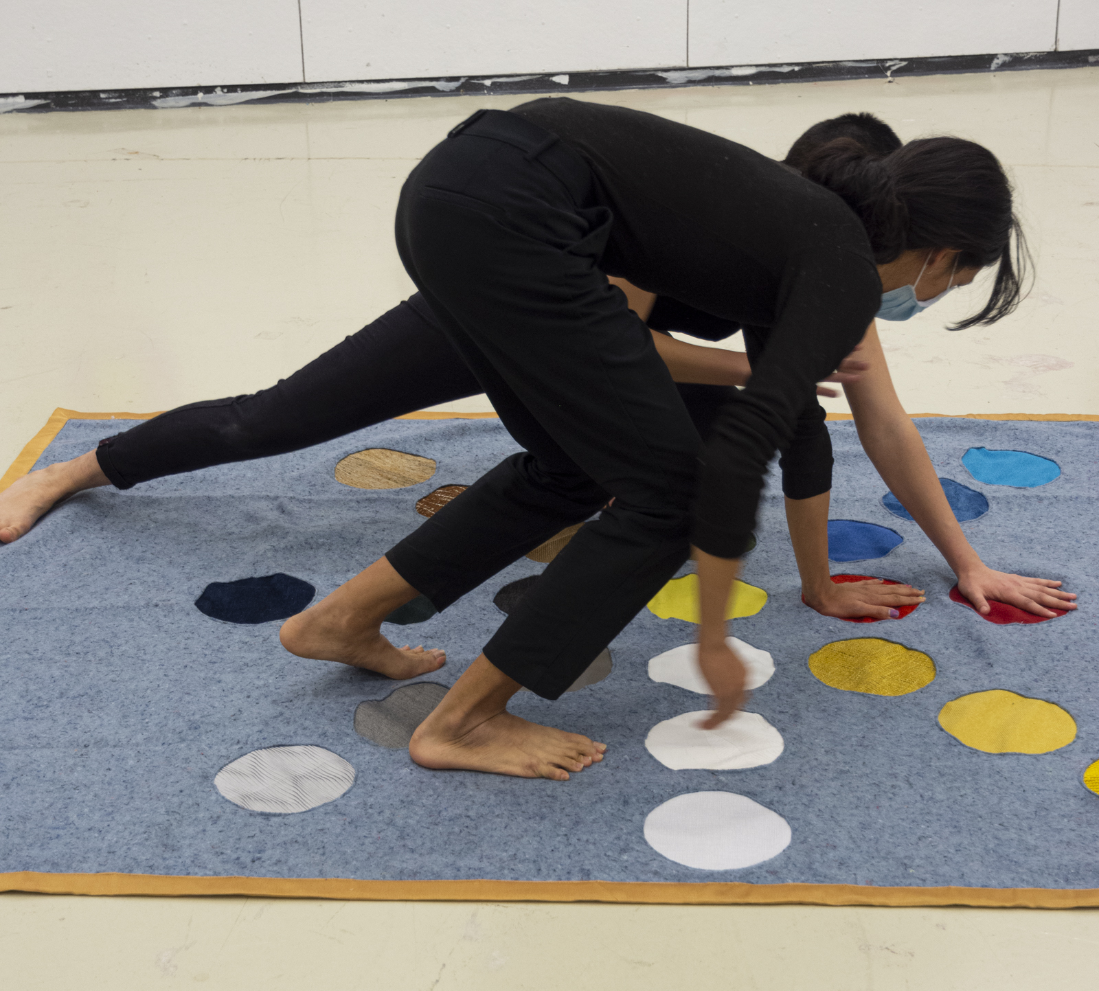 Two people wearing all black with all of their hands and feet touching the ground as if playing the game of twister. The mat they are playing on has colorful circles arranged to align in the 8 cardinal directions.