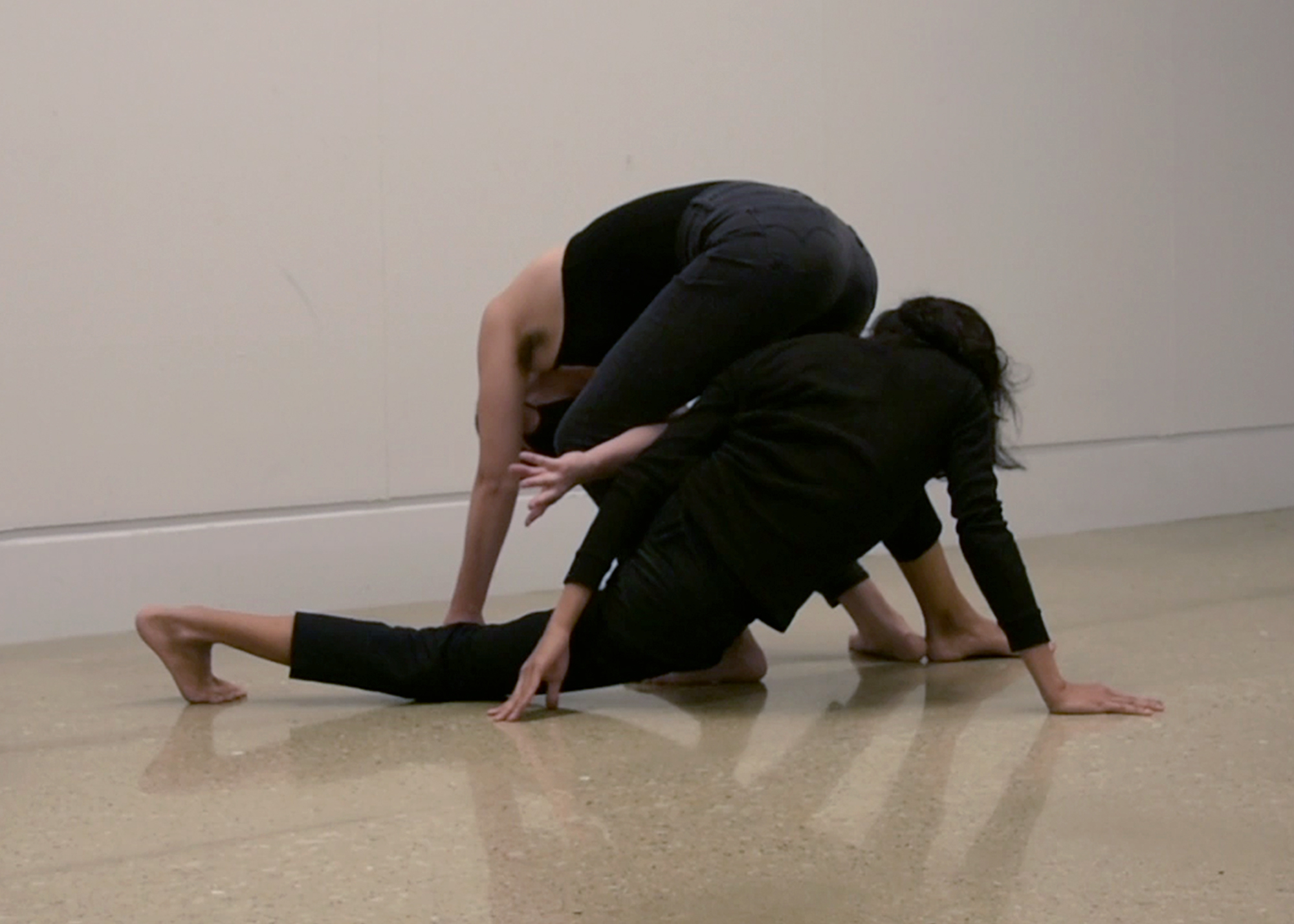 Two people dressed entirely in black entangled with each other. One of them reaches an arm between their legs, the other has all limbs touching the floor.