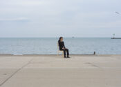 The artist is photographed from a distance wearing all black and seated on a wooden chair. Their face looks towards the right side. They are facing south. There is a lake behind them.