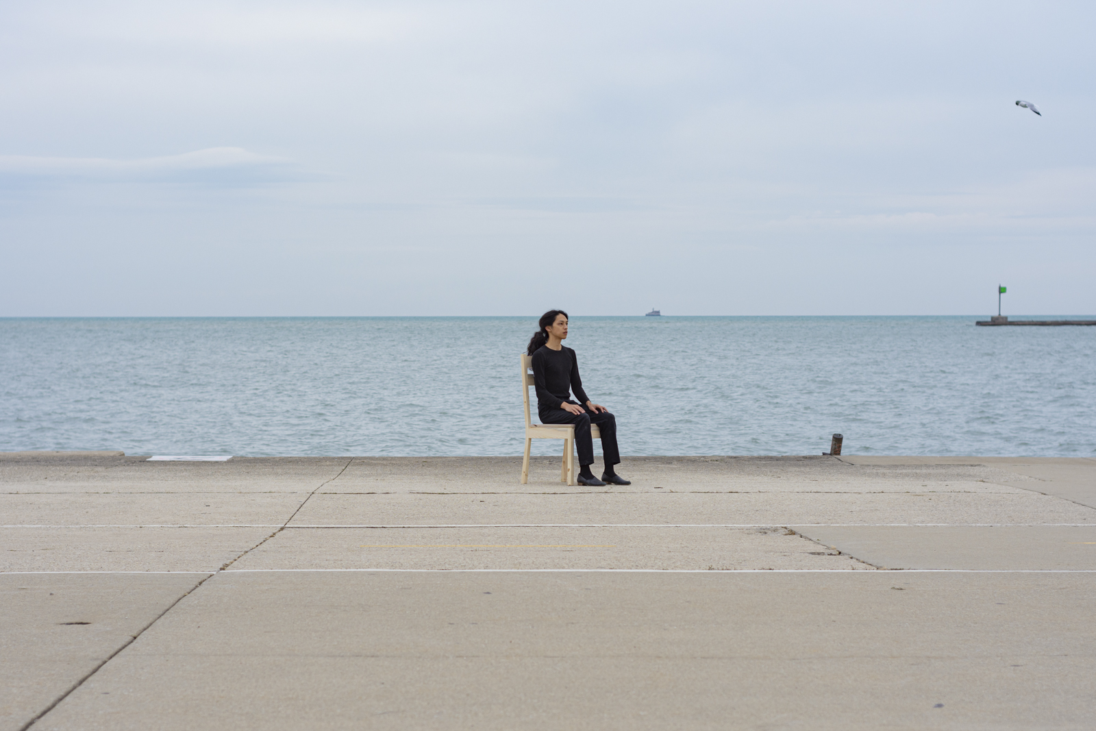 The artist is photographed from a distance wearing all black and seated on a wooden chair. Their face looks towards the right side. They are facing south. There is a lake behind them.