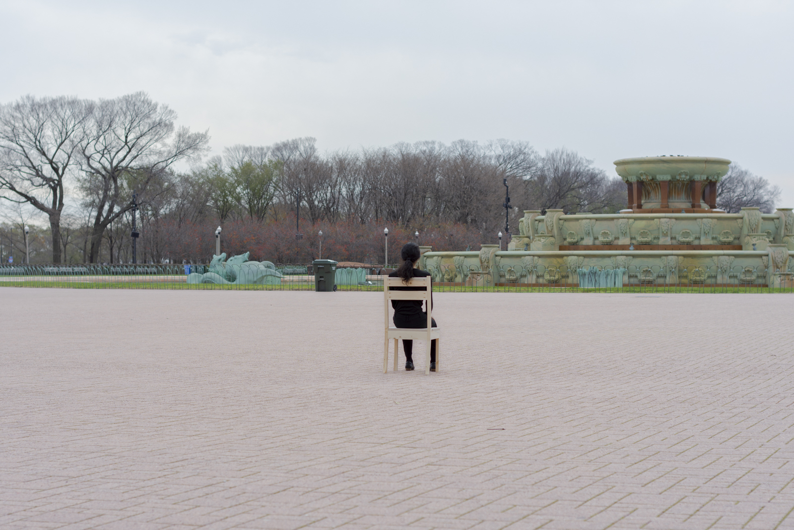 The artist is photographed from a distance wearing all black and seated on a wooden chair. Their face looks away from the camera. They are facing south. There is a fountain in front of them.