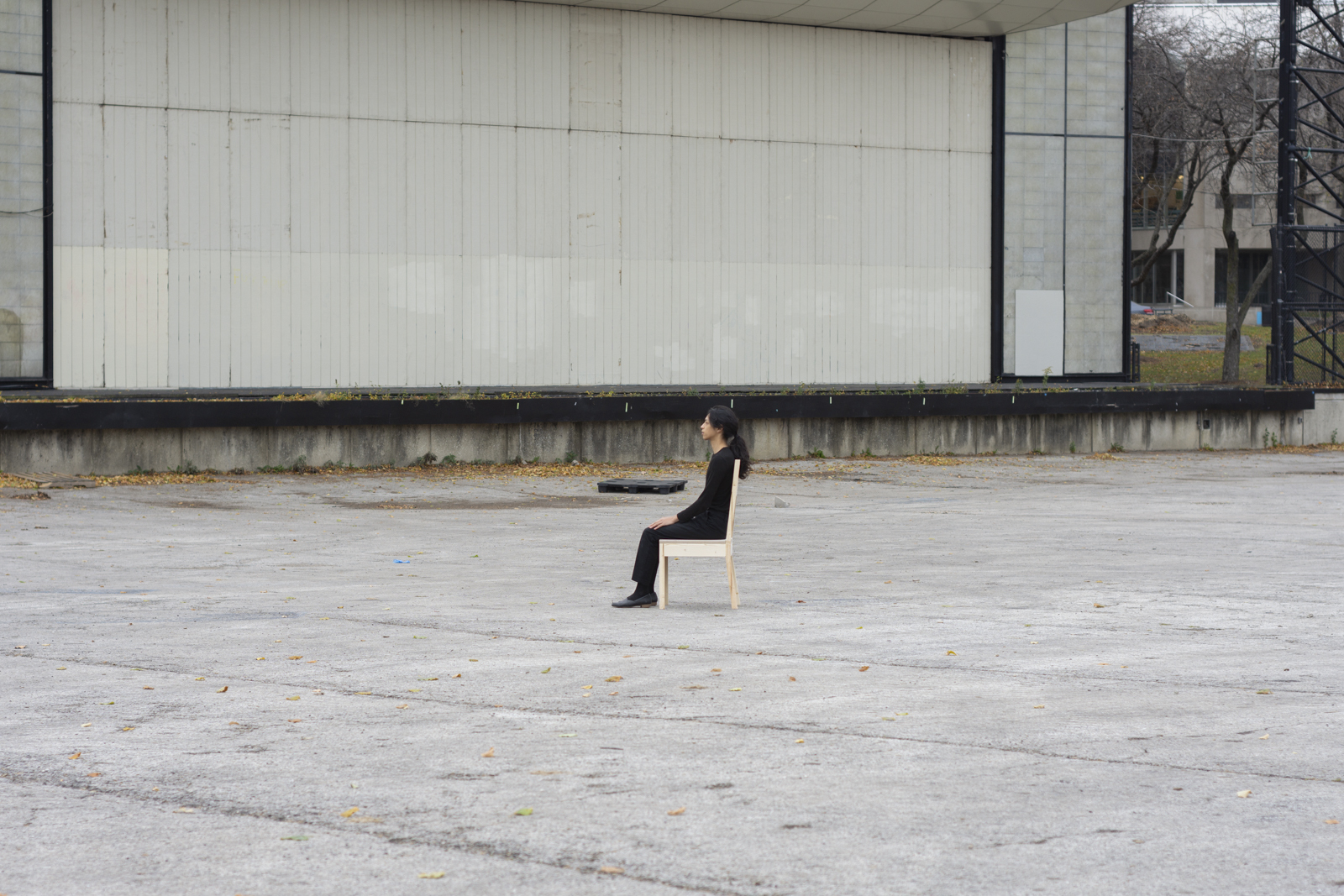 The artist is photographed from a distance wearing all black and seated on a wooden chair. Their face looks towards the left side. They are facing south. There is an amphitheater behind them.