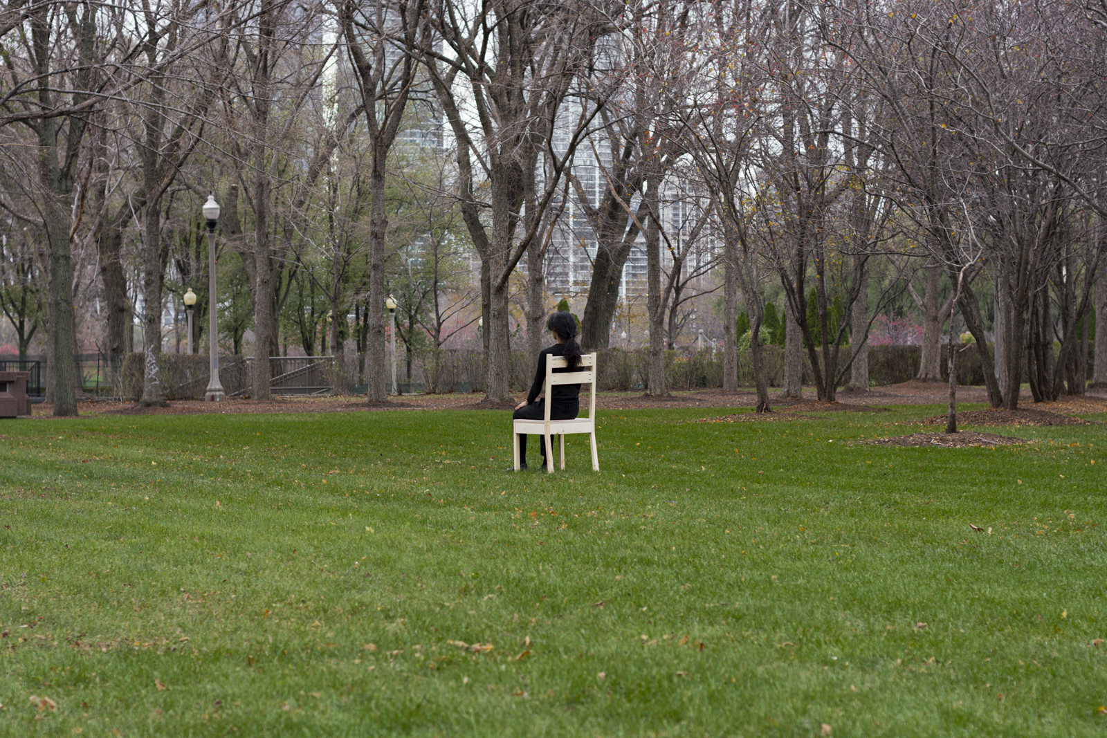 The artist is photographed from a distance wearing all black and seated on a wooden chair. Their face looks away from the camera. They are facing south. They are seated within a grassy park.