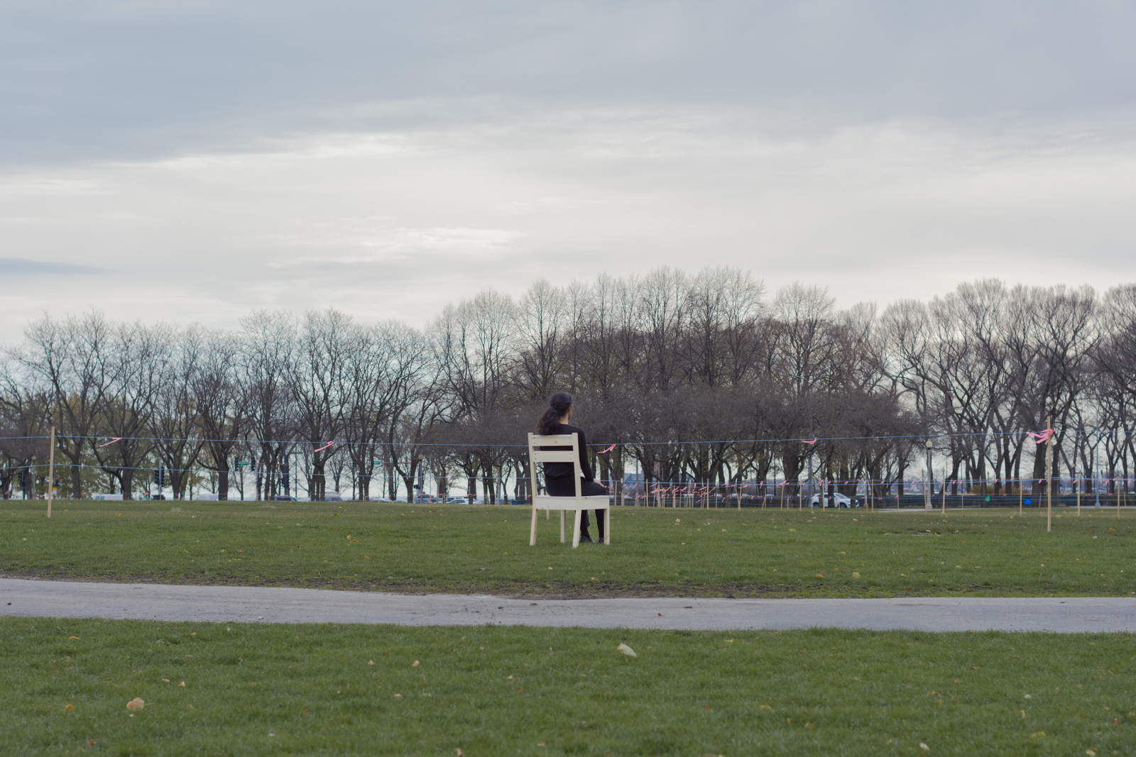 The artist is photographed from a distance wearing all black and seated on a wooden chair. Their face looks away from the camera. They are facing south. They are seated within a grassy park.