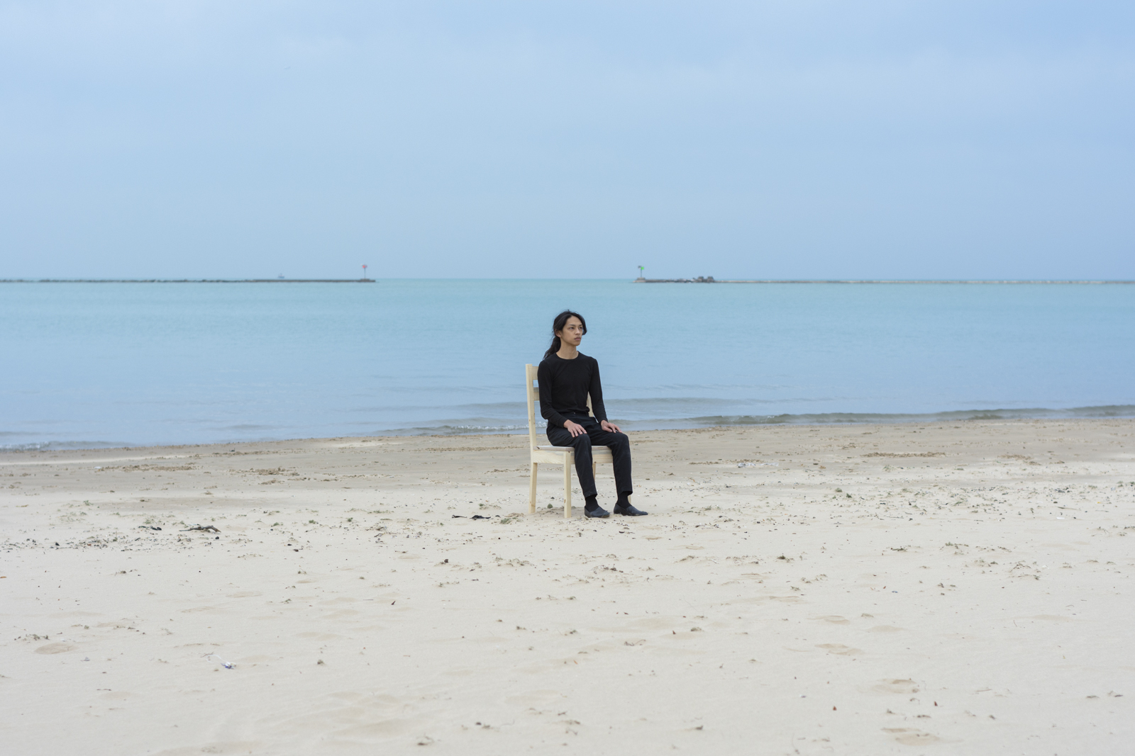 The artist is photographed from a distance wearing all black and seated on a wooden chair. Their face looks slightly to the right of camera. They are facing south. They are seated on top of sand at a beach.