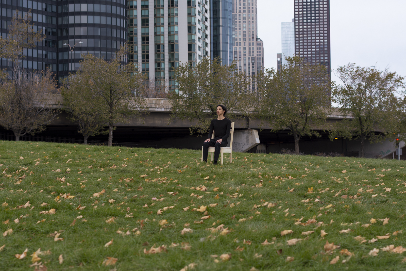 The artist is photographed from a distance wearing all black and seated on a wooden chair. Their face looks away from the camera. They are facing south. They are seated within a grassy park.