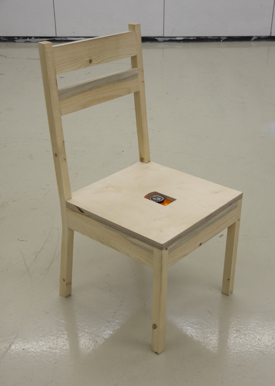 A simple wooden chair, with four legs and a backrest slightly angled back. In the seat of the chair is a specifically shaped divet that holds a compass.
