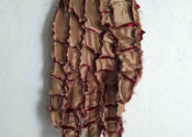 It is a golden color patchwork of fabric pieces formed into the shape of a hand with red fabric emerging from exposed seams. The hand is pointed downwards and mounted on a wall with the palm facing the viewer in varada mudra.