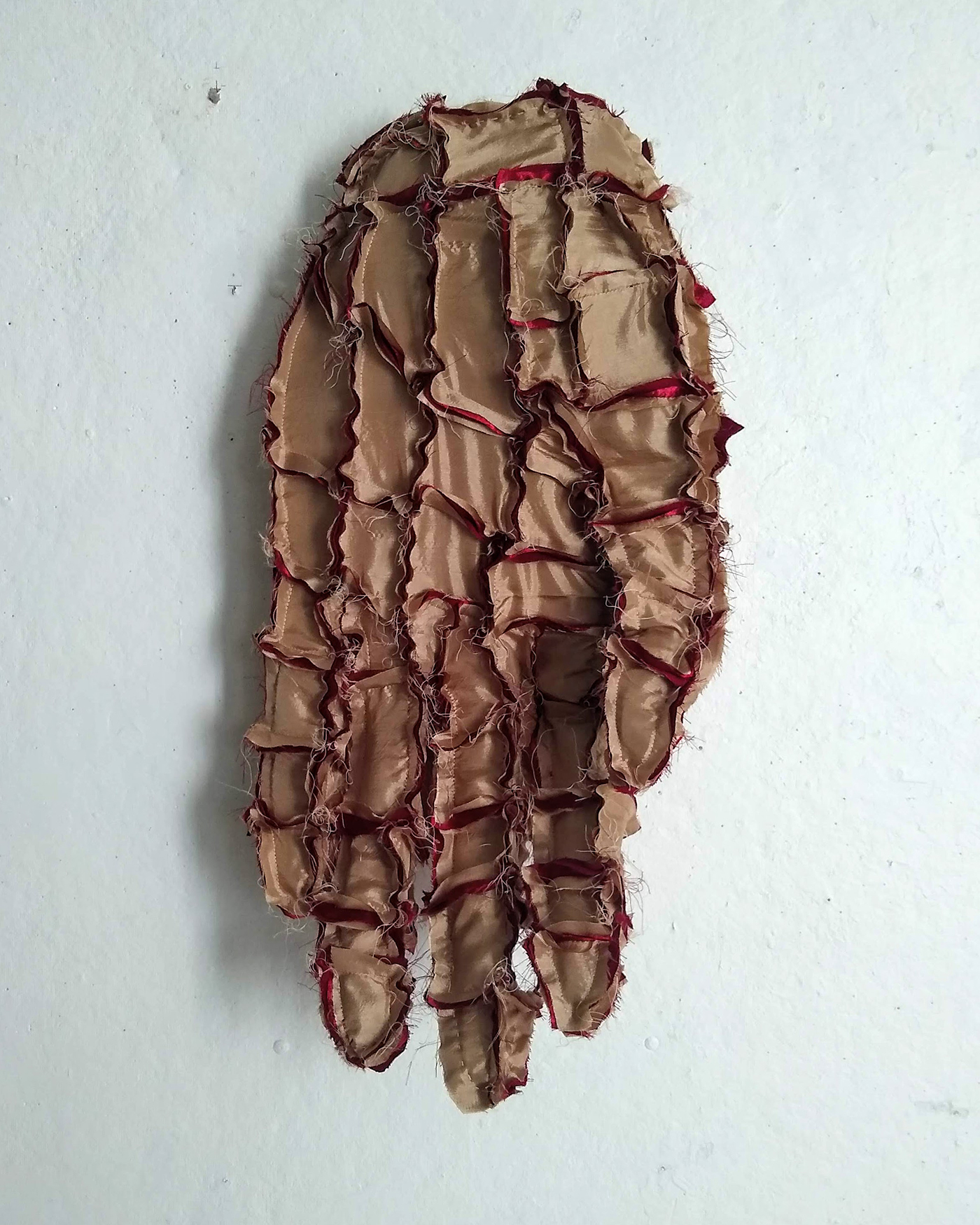 It is a golden color patchwork of fabric pieces formed into the shape of a hand with red fabric emerging from exposed seams. The hand is pointed downwards and mounted on a wall with the palm facing the viewer in varada mudra.