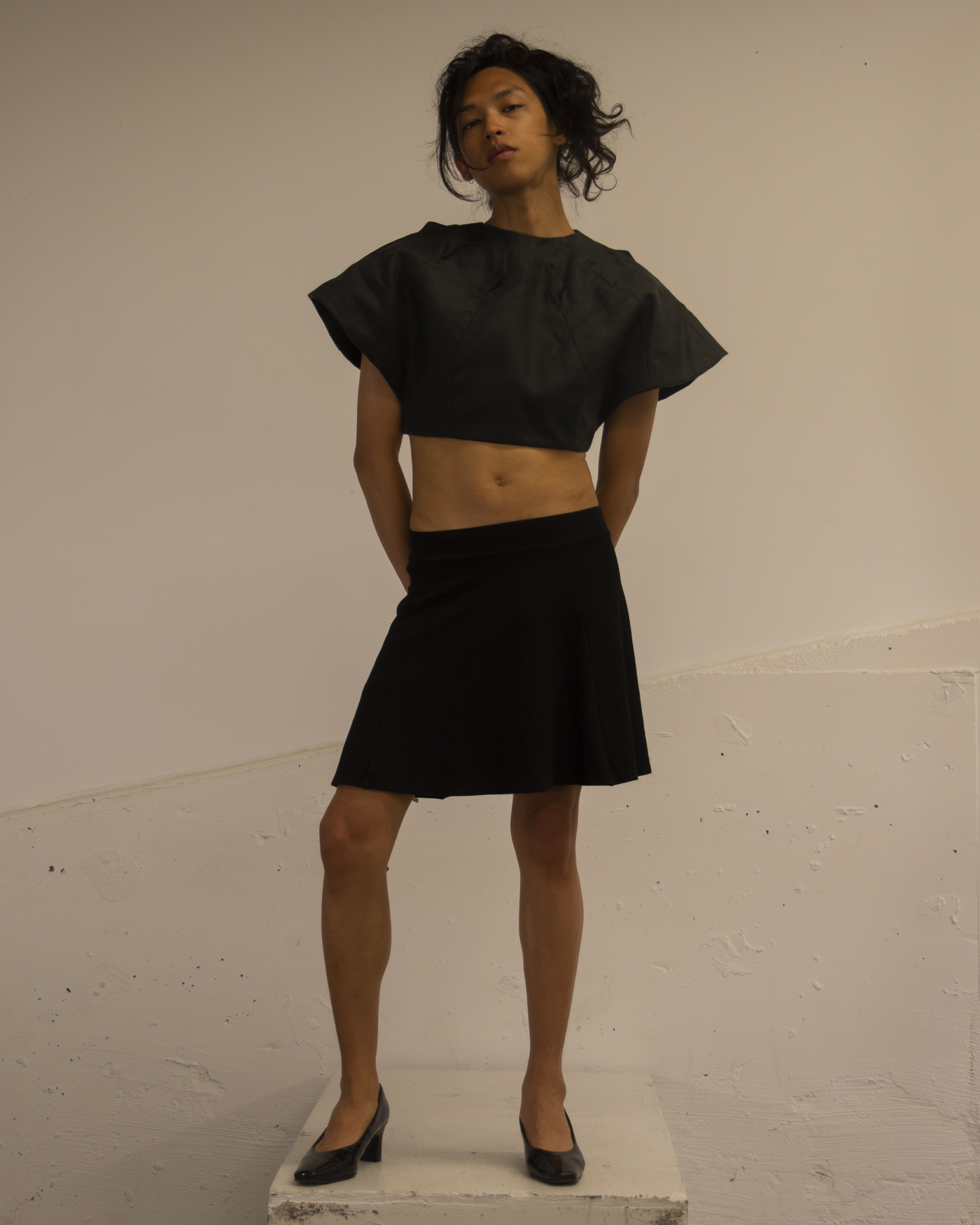 The Artist wears an angular top that is cropped at the waist along with a simple black skirt.