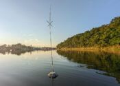 image of a buoyant steel shape in the middle of a lake that has what looks to be a ceramic candlestick holder tied on top of it. Stuck within this holder is a long reed with leaves tied at the top of it in the shape of an X