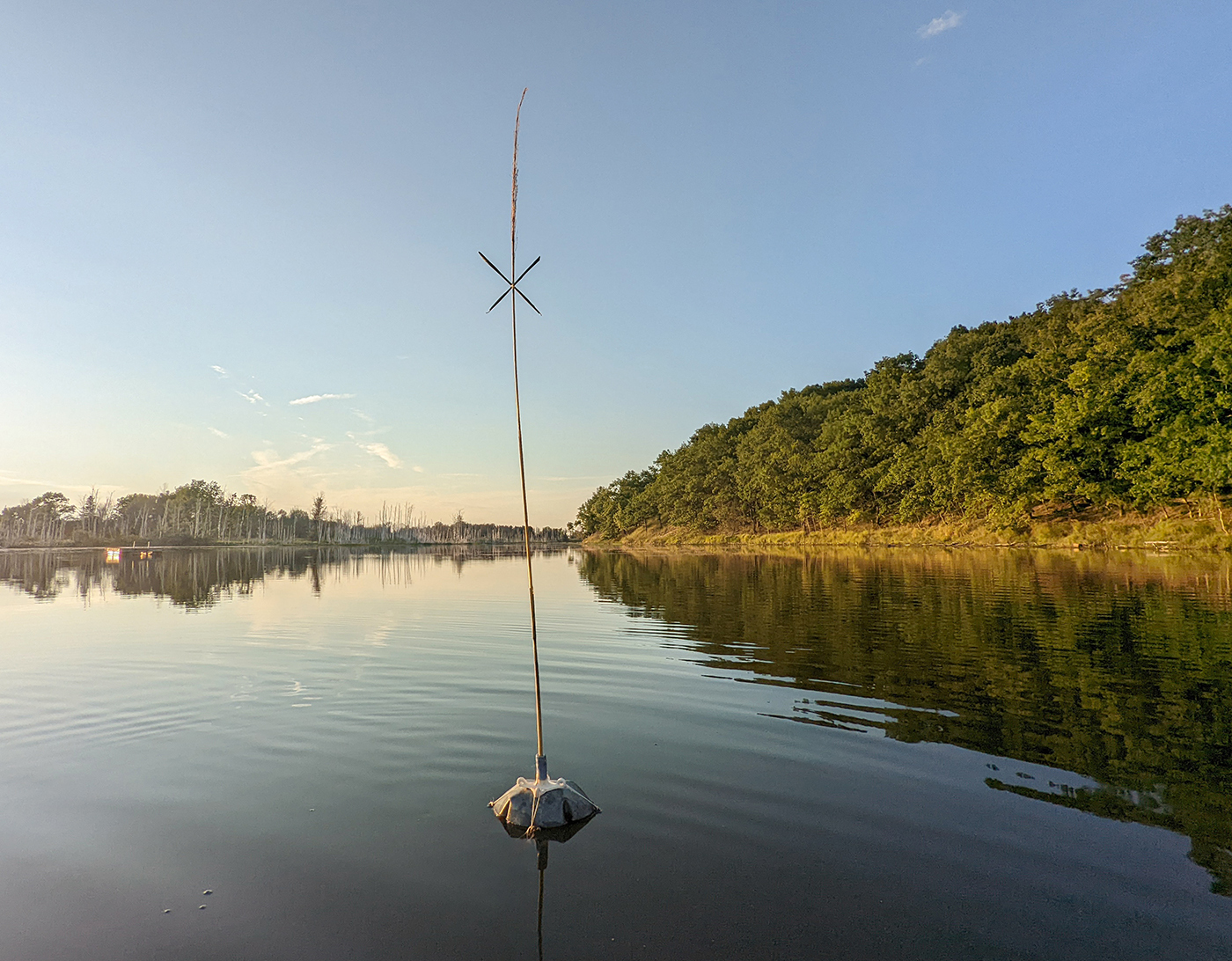 image of a buoyant steel shape in the middle of a lake that has what looks to be a ceramic candlestick holder tied on top of it. Stuck within this holder is a long reed with leaves tied at the top of it in the shape of an X