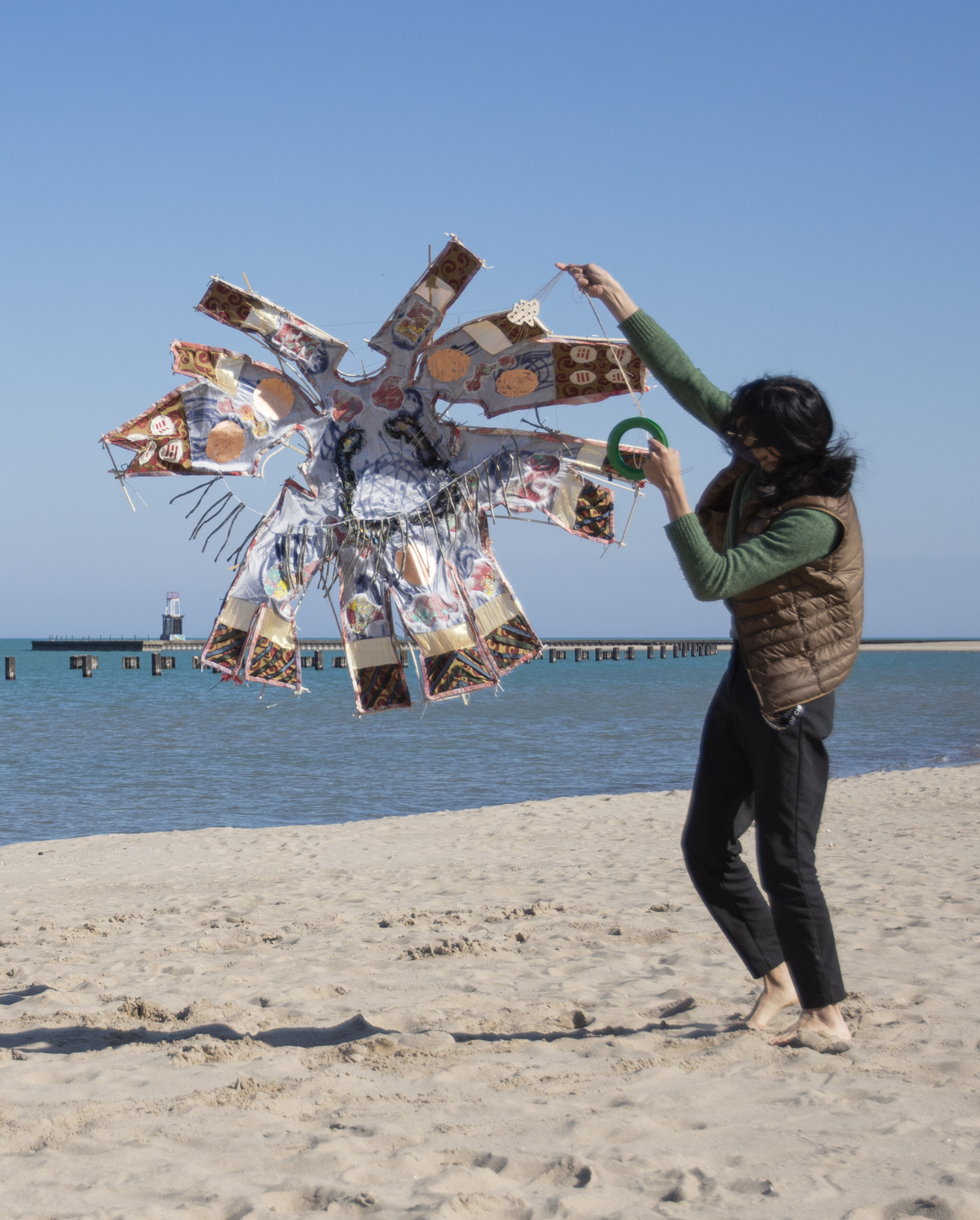 The artist is on the beach lifting their kite piece by its towline.