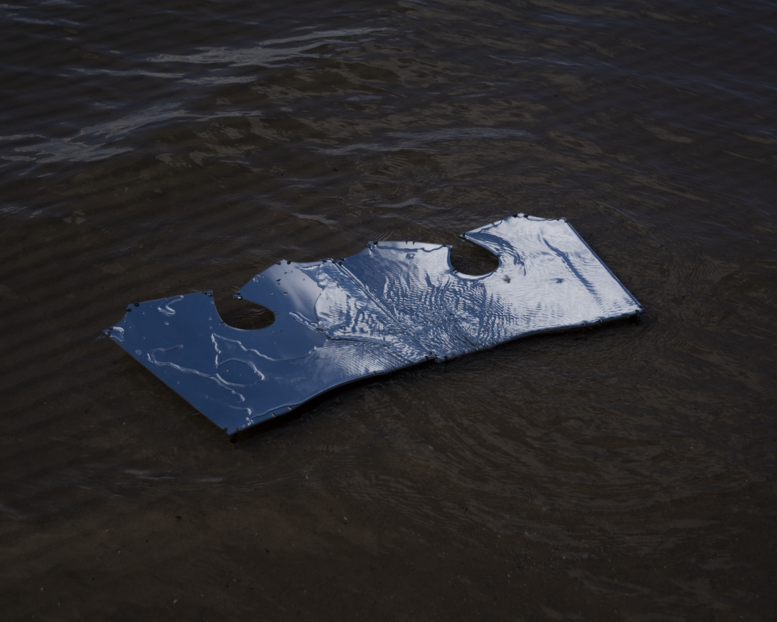 a set of mirrors in the shape of a bodice lies under some shallow water. Water laps over the mirror. The blue sky can been seen in it.