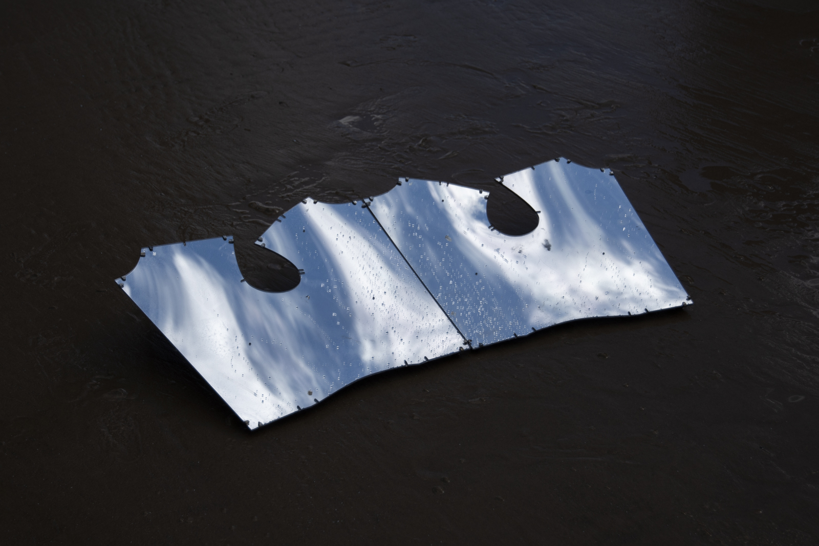 A wet mirror is angled on the beach such that the sky and clouds can be seen in its reflection. The wetness and shape of the mirror distorts the image.