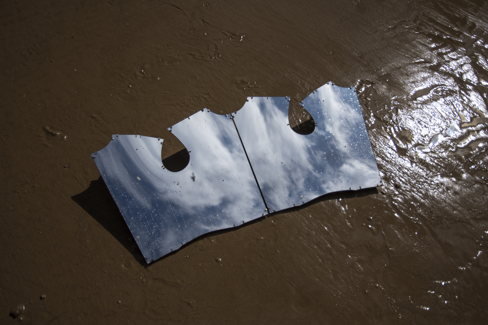 two mirrors in the shape of a bodice pattern are propped up on some brown sand. Clouds and blue sky are visible in the reflection.