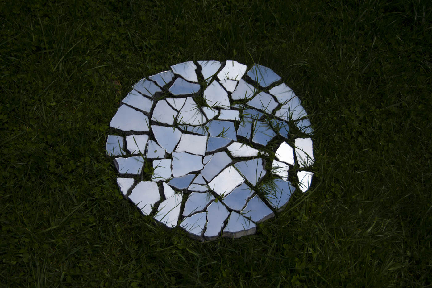 A circular shaped mirror is shattered and lying on the grass. Various shades of blue sky are reflected.